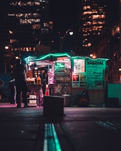 A vibrant street food stall illuminated by neon lights in a bustling city at night.